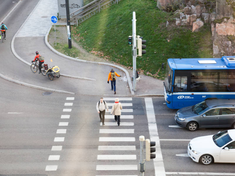 Jalankulkijoita ja pyöräilijöitä ylittämässä suojatietä. Risteyksessä pysähtyneenä bussi ja henkilöautoja.