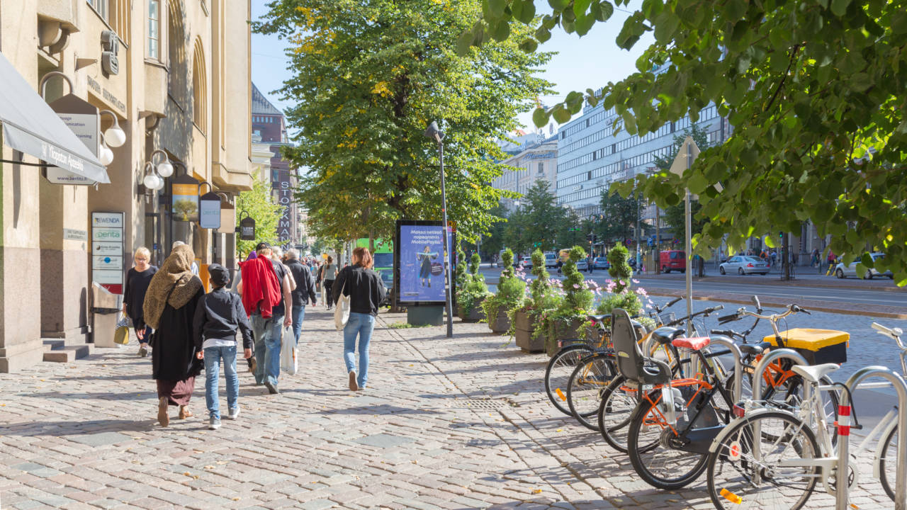 Jalankulkijoita Mannerheimintien jalkakäytävällä. Polkupyöriä parkissa, sivuilla taloja ja vihreitä puita.