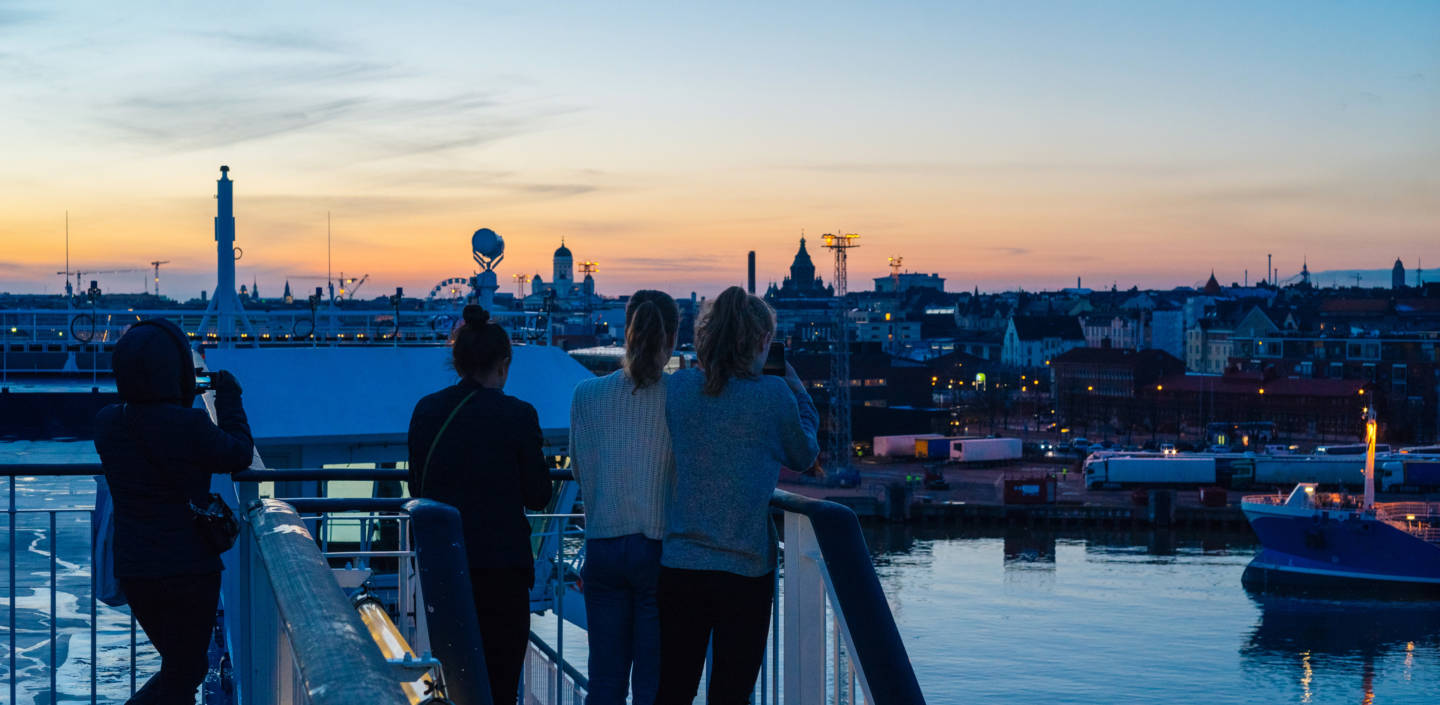 Neljä nuorta henkilöä ottavat kuvia Helsingistä laivan kannelta auringonlaskussa.