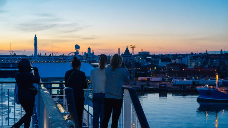 Neljä nuorta henkilöä ottavat kuvia Helsingistä laivan kannelta auringonlaskussa.