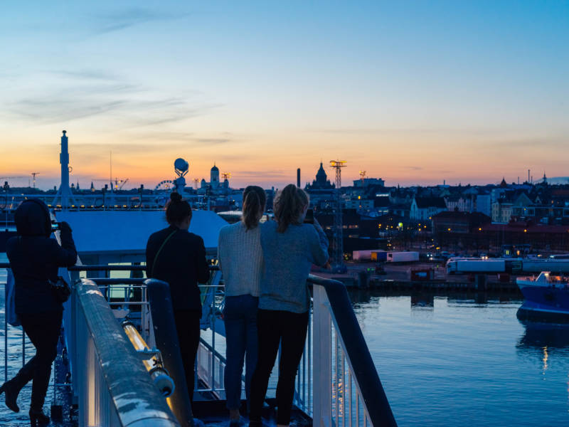 Neljä nuorta henkilöä ottavat kuvia Helsingistä laivan kannelta auringonlaskussa.