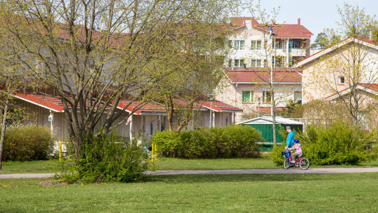 Keväinen puisto, taustalla taloja. Aikuinen kävelee ja lapsi pyöräilee.