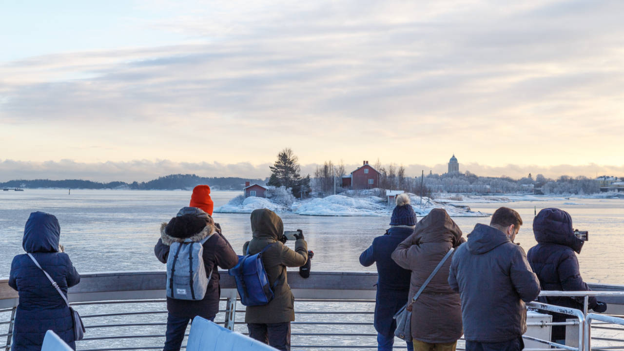 Ihmiset talvivaatteisiin pukeutuneena laivan kannella kuvia ottamassa. Meri ohuessa jäässä ja maassa lunta.