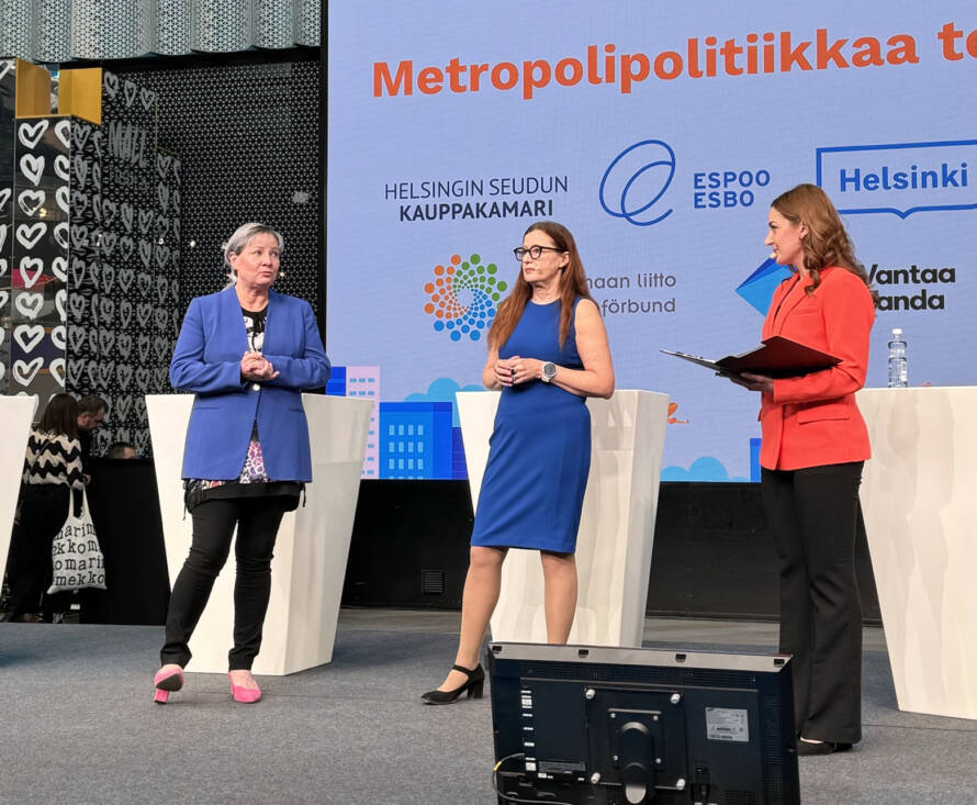 Kolme naista lavalla. Taustalla dia, jossa mm. Uudenmaan liiton, Helsingin seudun kauppakamarin ja pääkaupunkiseudun kaupunkien logot.