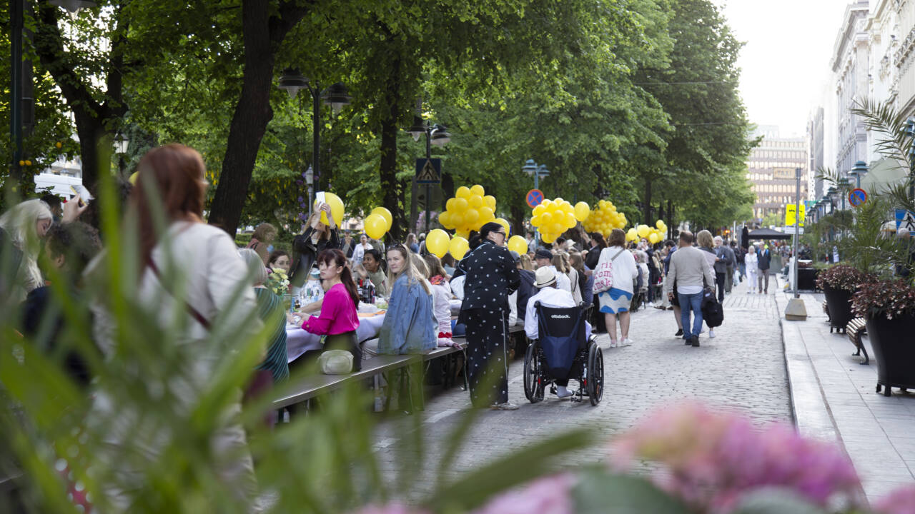 Ihmisiä Helsingin Esplanadilla pöydän ääressä ruokailemassa. Ilmapalloja pöydän äärellä.