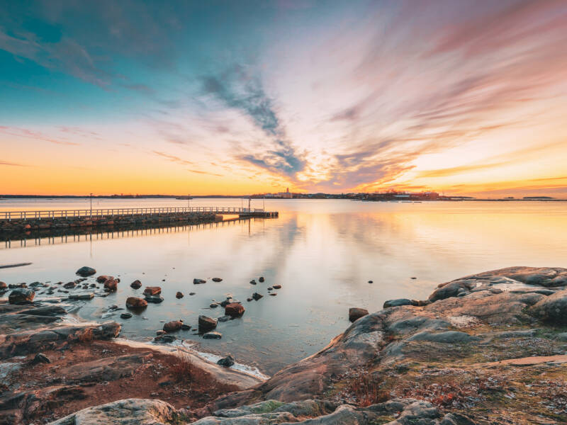 Tyyni meri, vasemmalla pitkä laituri, etualalla kallioista rantaa. Kauempana merellä Suomenlinna.