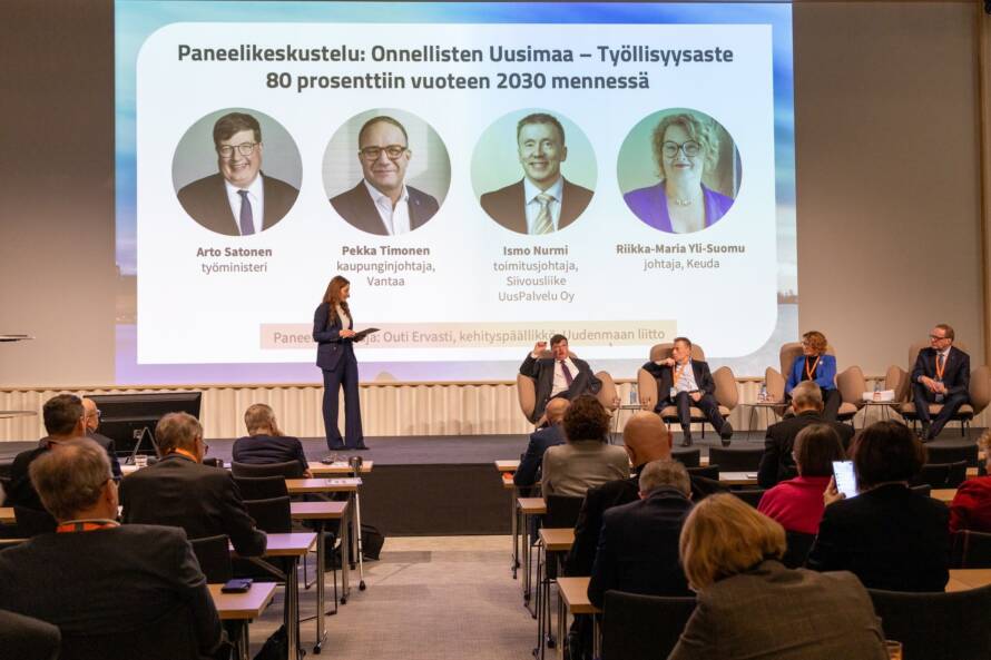 Kokoustilan takaa otettu kuva, jossa näkyy yleisöä ja lavalla neljä tuoleissa istuvaa panelistia ja seisova juontaja. Takana valkokangas, jossa panelistien nimet ja kuvat.