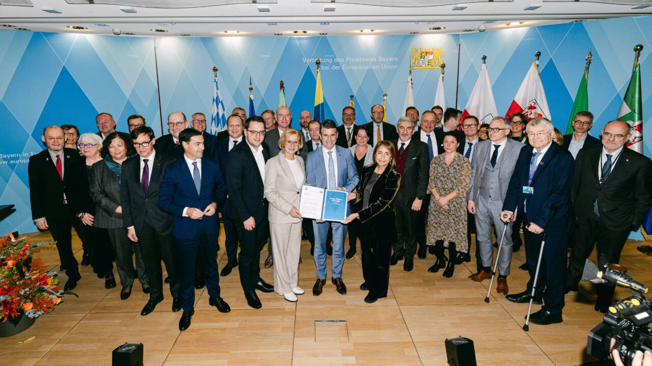 Participants of the Power Regions of Europe meeting standing on stage in front of the blue wall and national flags. The three people standing in front are holding a signed declaration. A flower arrangement is on the left of the stage and a system camera is visible on the right.