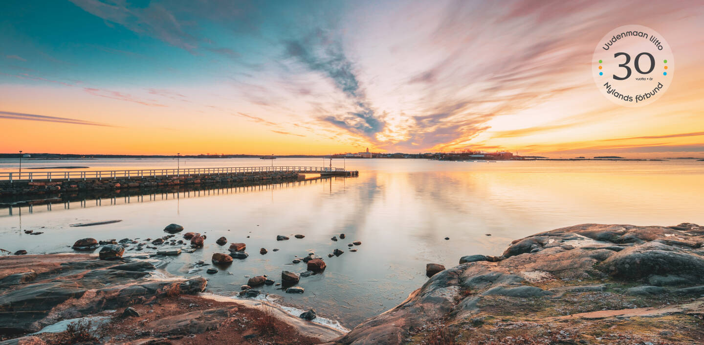 Tyyni meri auringonlaskussa. Rannassa kallioita ja laituri merelle. Logo Uudenmaan liitto 30 vuotta.