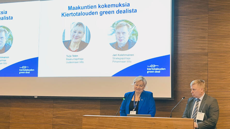 Kaksi henkilöä seisomassa puhujapöntössä, jonka takana on valkokangas, jolle on heijastettu puhujien kuvat ja teksti Maakuntien kokemuksia Kiertotalouden green dealista